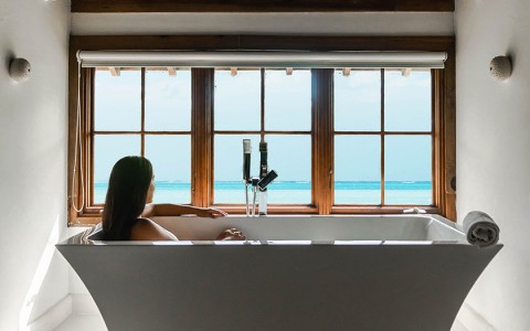 A woman sitting in an oversized bath tub looking out at the view of the bright blue water with dark oak ceilings above.