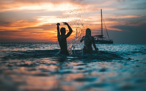 Two people splashing water at each other in the ocean while the sun is setting.