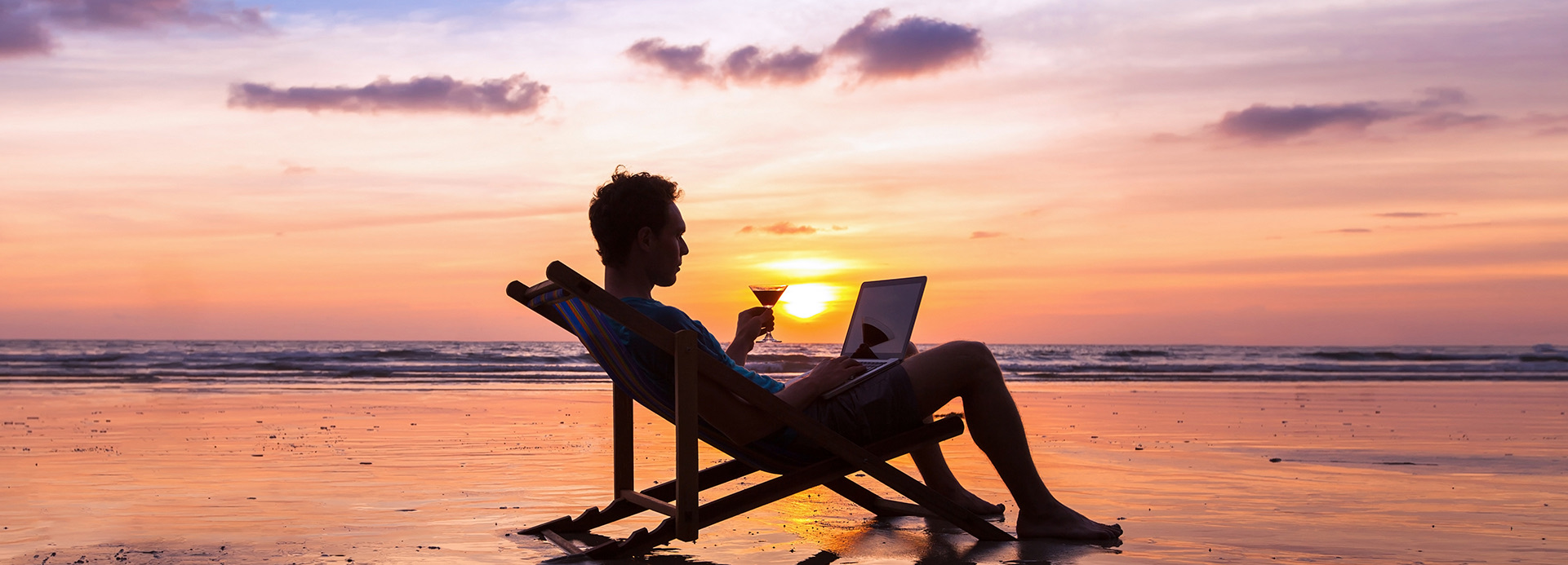 A man sitting in a lounge chair sipping a cocktail with his laptop at sunset.