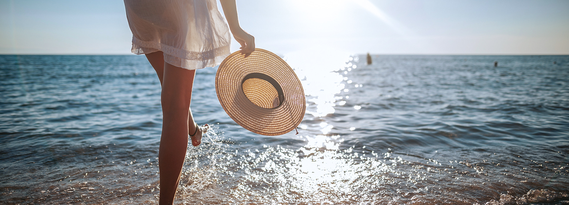 Woman walking into the ocean wearing a white coverup and holding a tan straw hat at sunset.