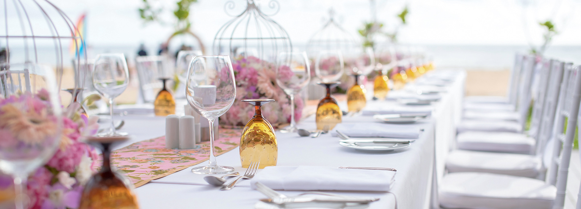 Long white linen covered table setup for dining on the beach during the day.