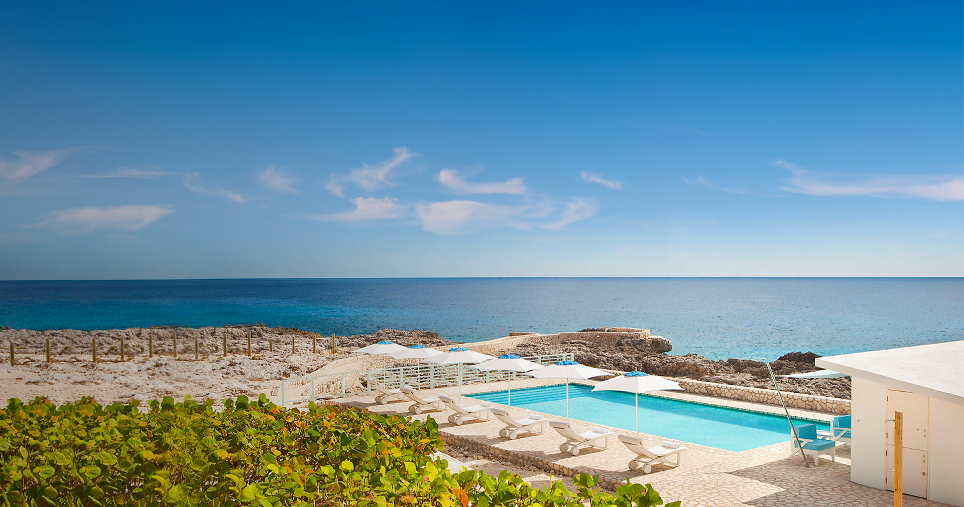 Crystal clear pool located next to the ocean and white lounge chairs with opened umbrellas.