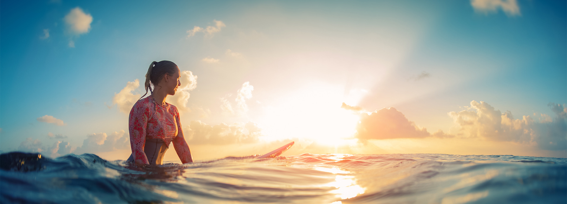 Woman sitting on surfboard looking off into the bright sunset.