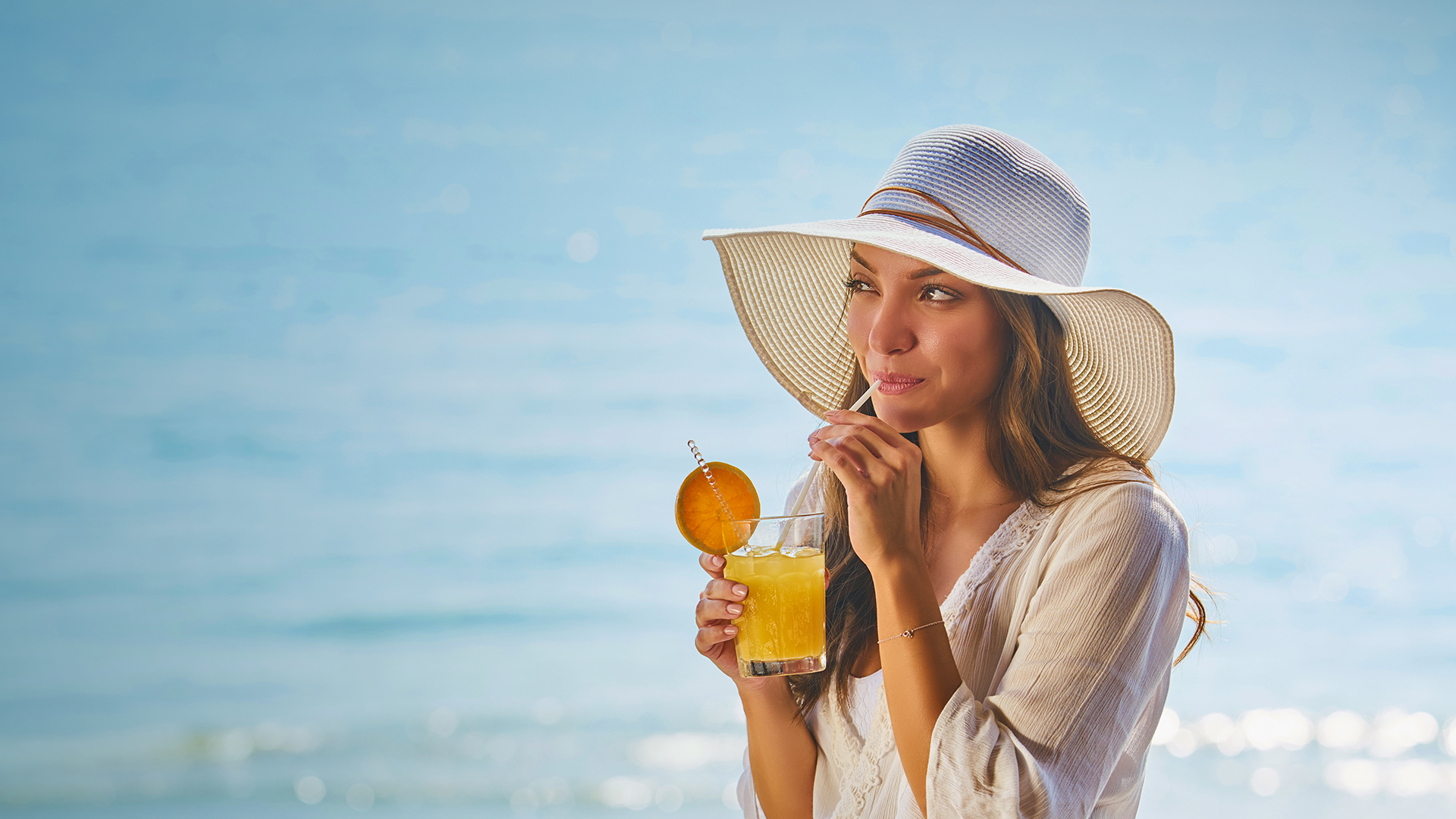 A woman wearing a light tan straw hat sipping an orange drink at the beach during the day.
