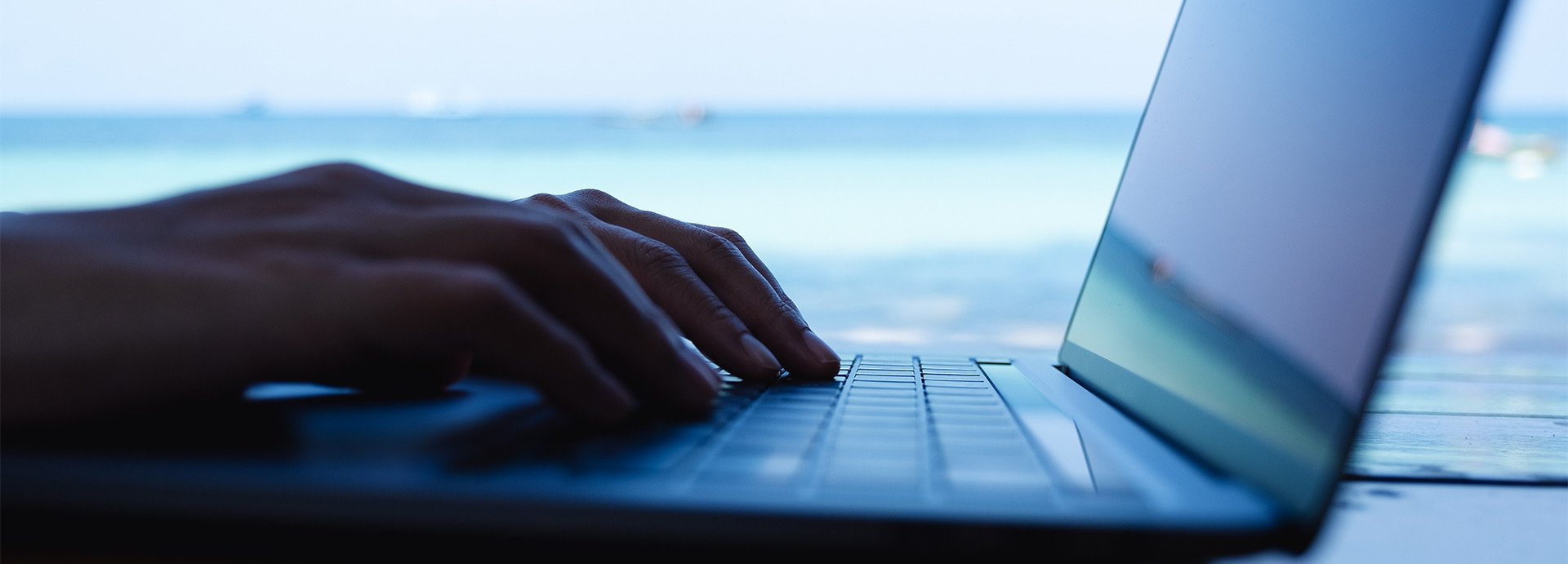 A person typing on an opened laptop next to the ocean during the day.