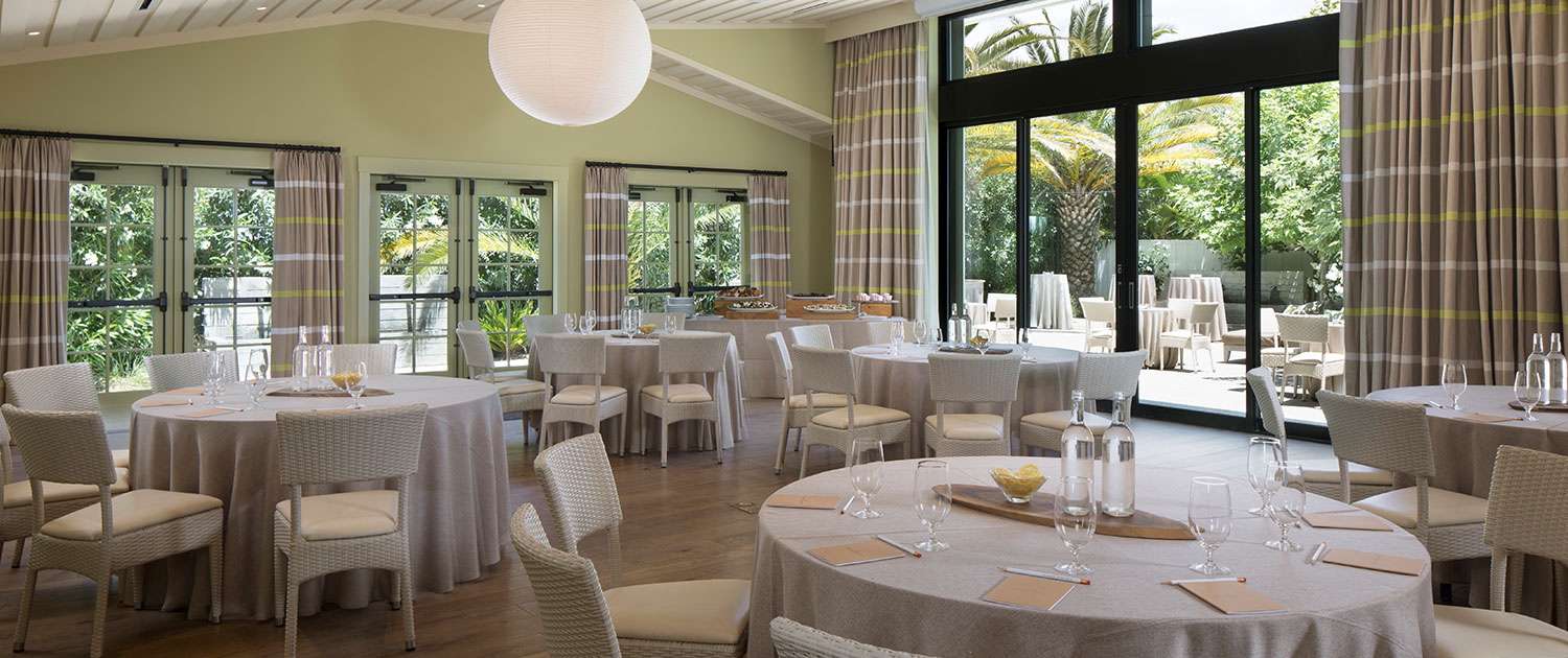 room filled with round tables draped in light gray tablecloths with large windows and doors open to an outdoor area