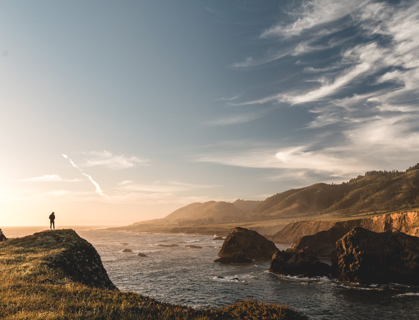 a wide shot of a person in the far distance standing on a cliff overlooking the ocean at sunrise