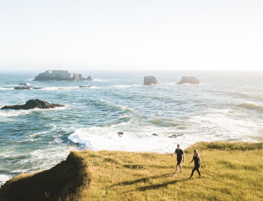 man and woman walking towards the edge of the grassy cliffs to look at the ocean water