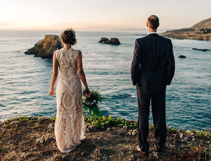 bride and groom standing on rocks overlooking the ocean