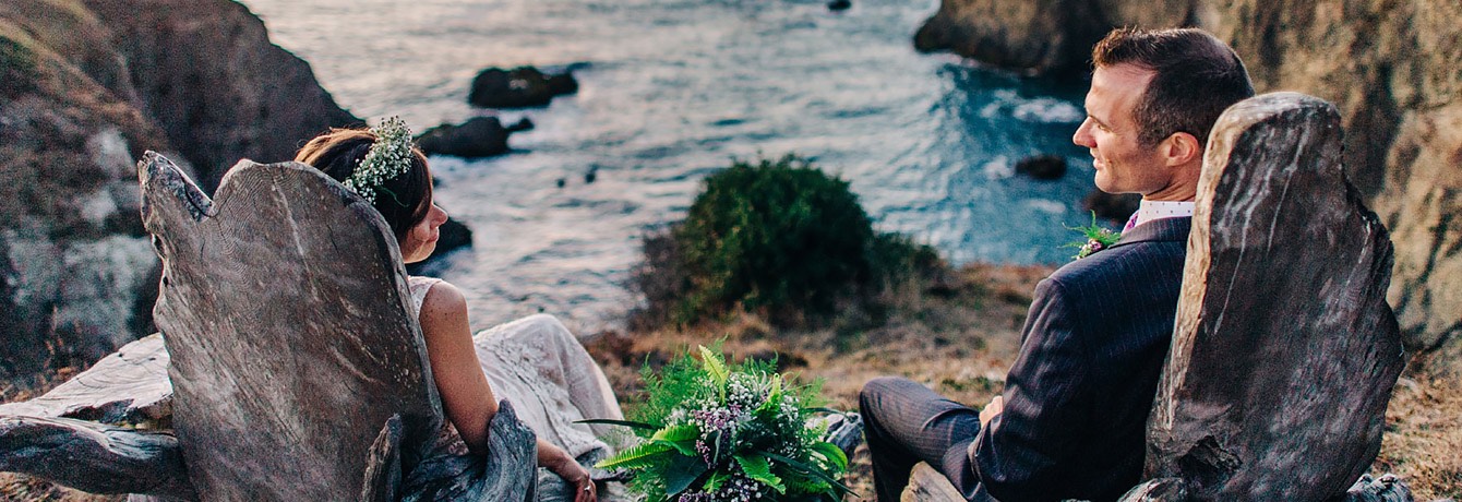 bride and groom sitting on wooden lawn chairs overlooking the ocean from the cliffs
