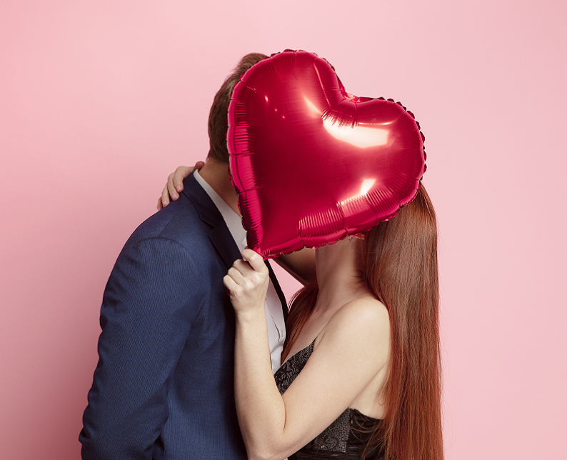couple in front of pink wall with a big red heart balloon covering their faces while they kiss