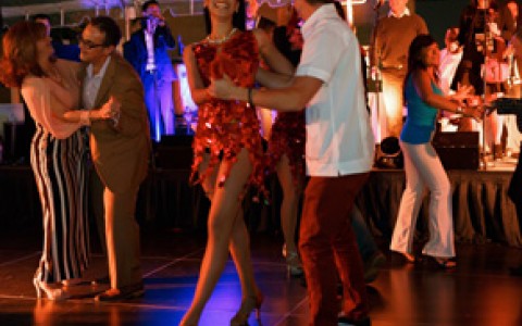 People dancing salsa, main focus is a girl wearing a red dress dancing with man in a white shirt and straw hat