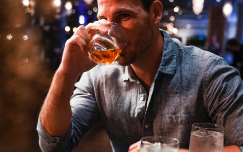 smiling man taking a sip of his drink while sitting at a table with food 