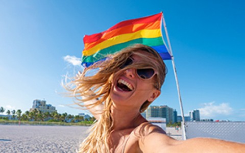 a woman taking a selfie on the beach with the pride flag blowing in the wind behind her