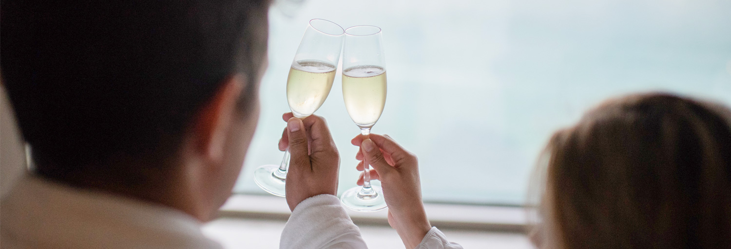 a couple holding up champagne glasses and doing a cheers with the water view from their guestroom