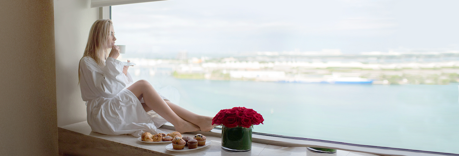Woman sipping coffee in her robe, overlooking the water in Miami from her hotel room view