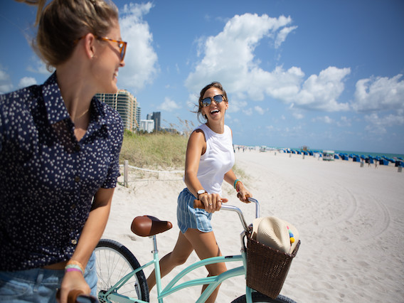 two women walking their bicycles onto the beach and laughing