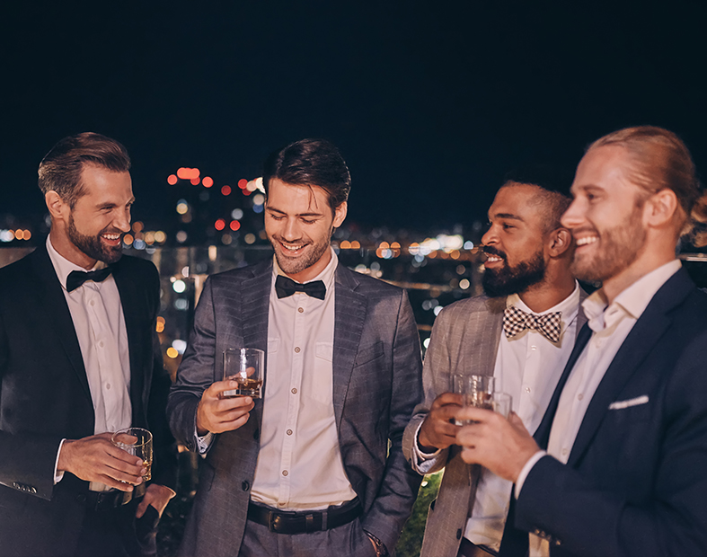 Four males smiling and holding cocktail glasses in their hands on an outdoor terrace.