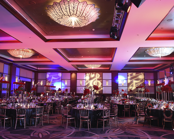 A dim lit ballroom with tables set for dinner and red floral centerpieces.