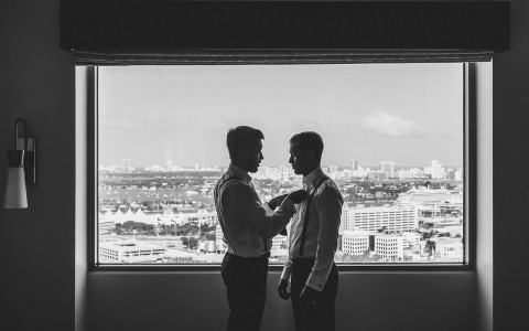 A man helping another man put on his tie with the view of the city outside the window.