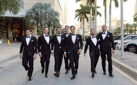A group of seven men wearing black tuxedos while smiling and walking down the street at sunset.