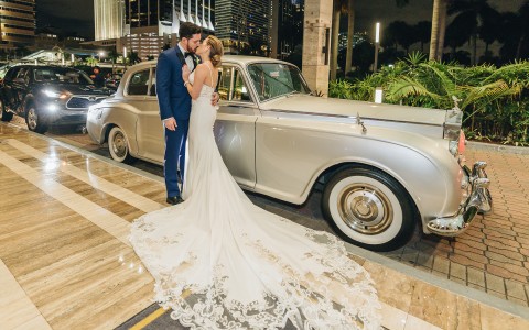 A bride and groom kissing next to an old fashioned silver car.