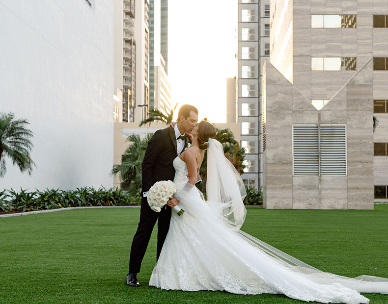 A bride and groom kissing on a lawn with the sun setting.