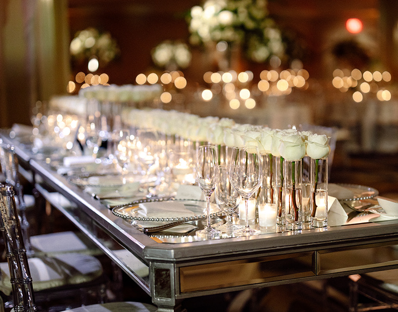 Long table with multiple white roses as the centerpieces and soft lighting from candles.