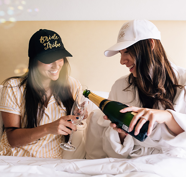 Two girls wearing baseball caps and pouring champagne.