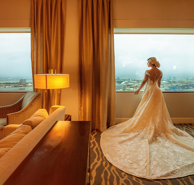A bride in a dim lit room looking out the window at the view of the city.