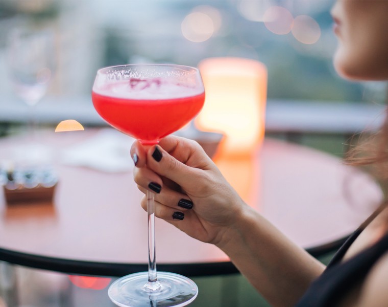A woman holding a pink cocktail at a small round table.