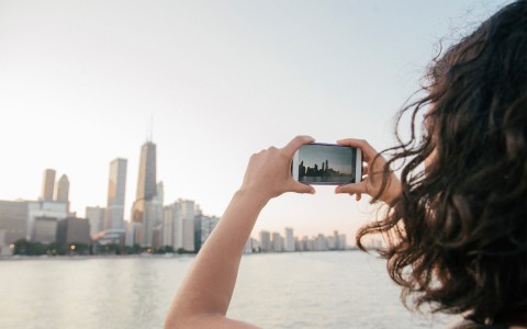A woman talking a picture of the buildings with her cellphone at sunset.