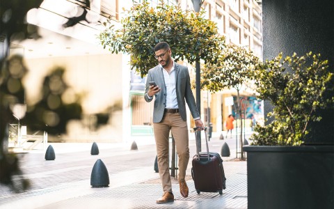 A businessman walking down the sidewalk with his phone in one hand and his luggage in the other hand.