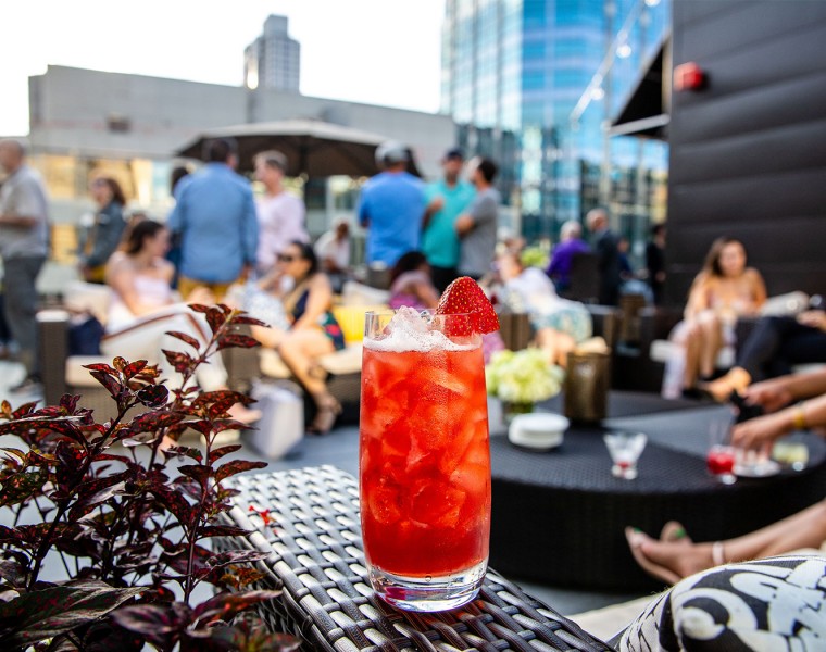 A pink beverage served with fruit and lots of people relaxing the background on a rooftop terrace.