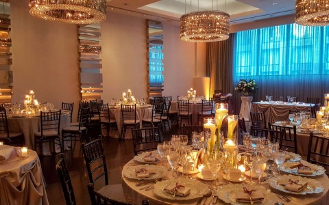 A low lit dining room with chandeliers hanging above and lots of tables set for dinner.