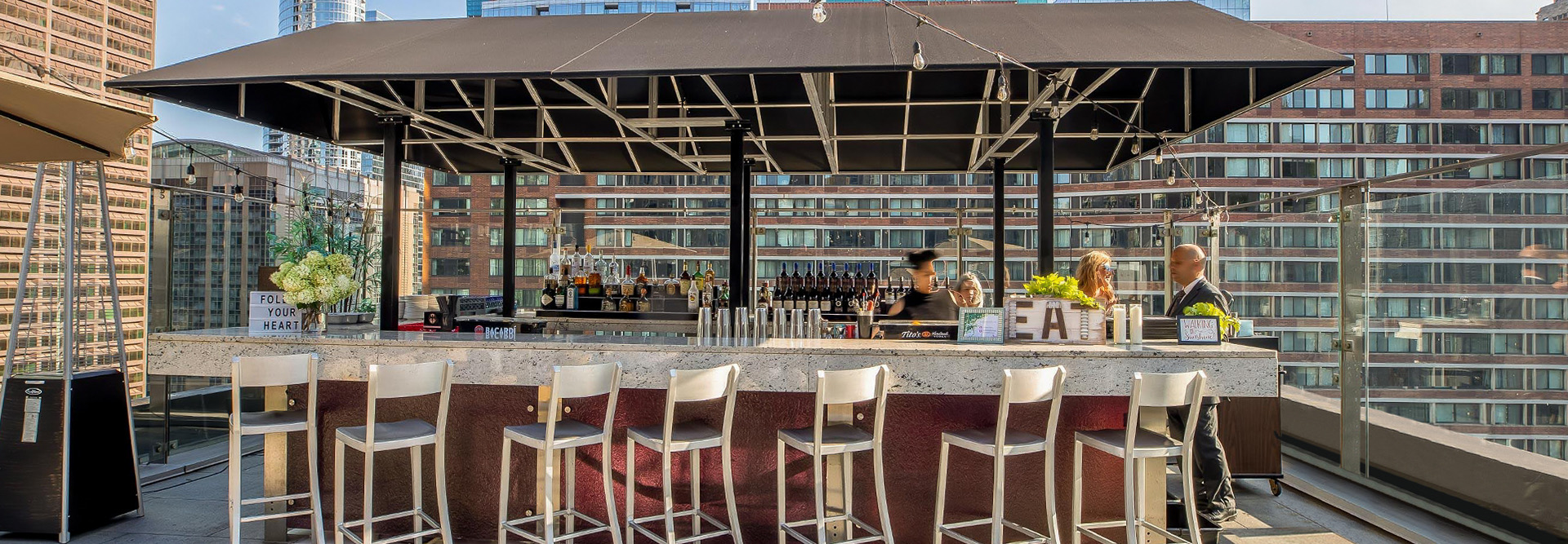 An outdoor bar and stools on a rooftop terrace with people sitting at it.
