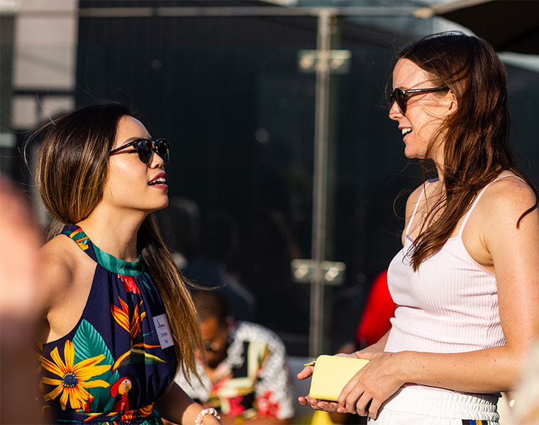 Two woman wearing sunglasses and talking to each other outside with the sun shining.