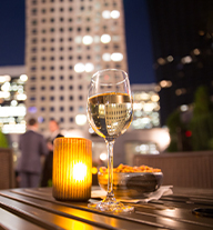 A glass of white wine on a table next to a candle and a tall building in the background.