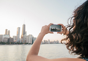 A woman holding her phone taking a picture of the buildings and the sun setting in the distance.