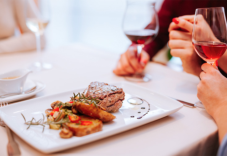 People drinking red wine at a table with steak served on a white plate.