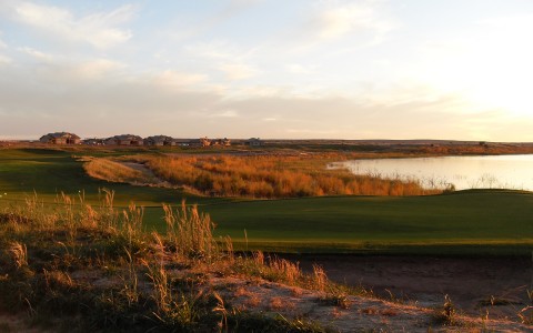 the sun setting over the lake and golf course with tall grass along the shore