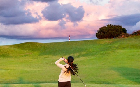a woman finishing her golf swing on the course with the sun setting