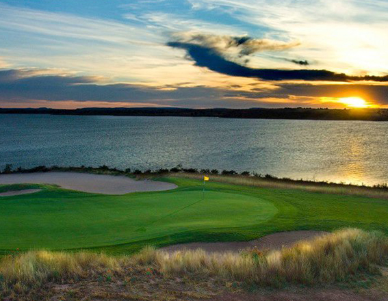 tall grass and a bright green course next to a large body of water as the sun is setting