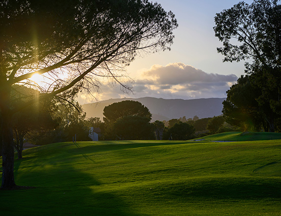 the sun setting to the left of the golf course with dark clouds over the mountains