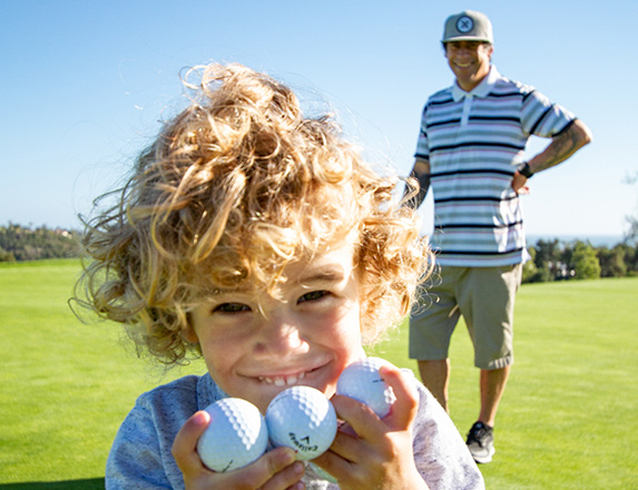 a small kid holding three golf balls in his hands and smiling with a man standing just behind him on the golf course