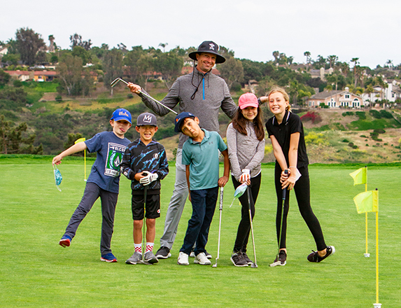 a group of kids and an instructor with golf clubs in their hands posing for a photo on the golf course