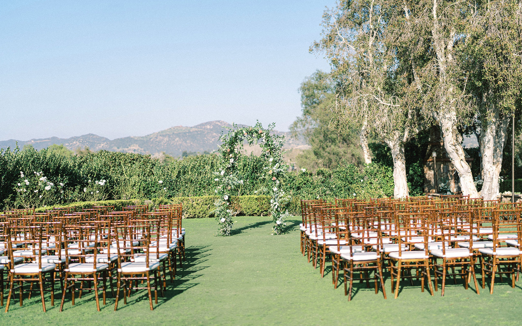 an area set up for an outdoor wedding with wooden chairs and a floral arch