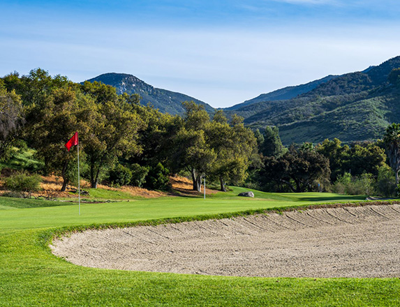 a red flagstick and a large sand dune with mountains in the distance and clear blue skies above