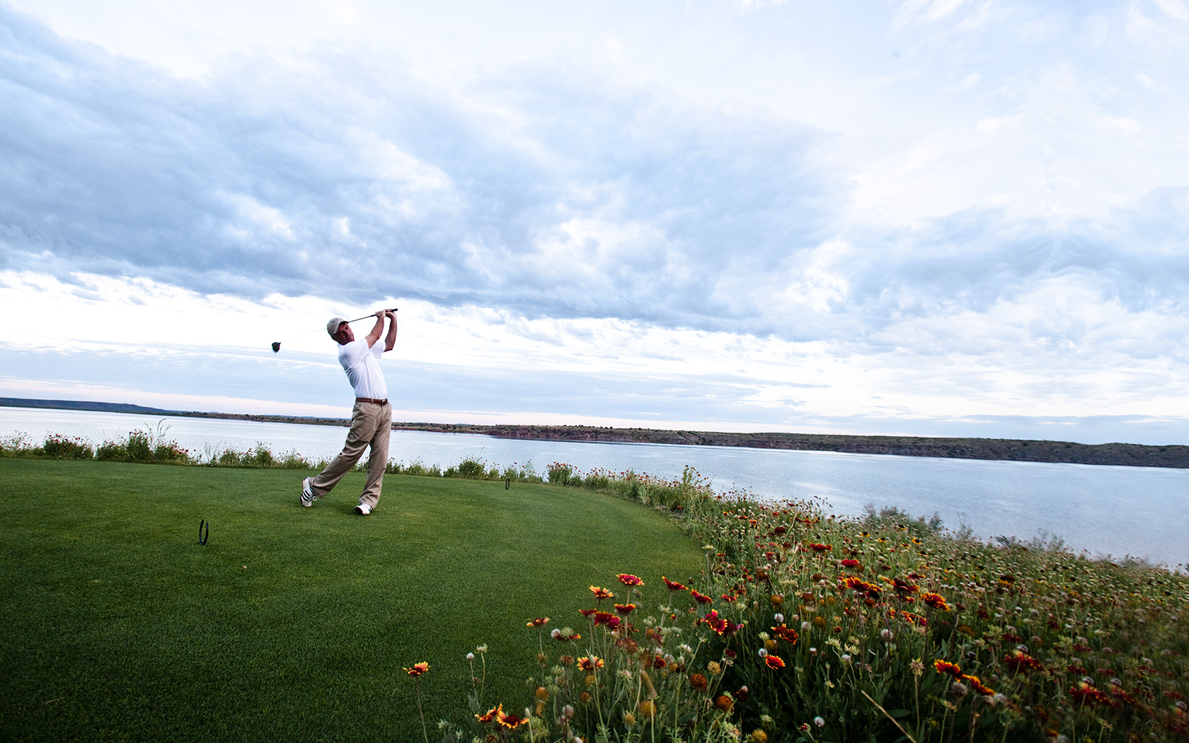 a man finishing his swing on the putting green located near a large body of water