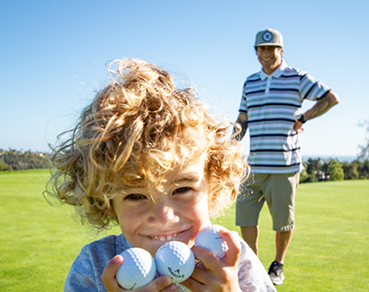 a young little boy smiling with three golf balls in his hands and an older man standing behind him